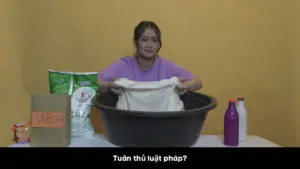 Young woman washing clothes in a large tub with detergent and fabric softener on a table against a yellow background.