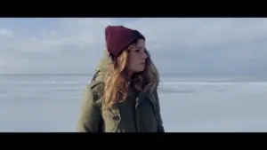 Woman in a winter coat and beanie stands on a snowy landscape, gazing thoughtfully at the horizon under a cloudy sky.