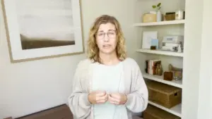 A woman with curly hair wearing a light sweater stands in a bright, modern room with shelves displaying decorative items.