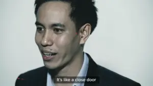 Smiling young man in a suit speaking to the camera, with a neutral background and subtitles at the bottom.