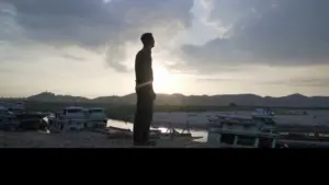 Silhouette of a person standing by a river at sunset, with boats and hills in the background, creating a serene atmosphere.