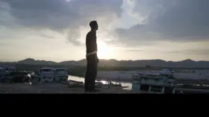 Silhouette of a person standing by the water at sunset, with boats and mountains in the background.