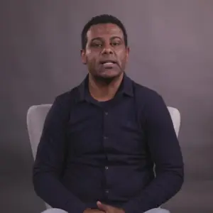 Man in a black shirt seated against a gray background, looking directly at the camera with a neutral expression.