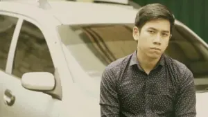 Young man in a patterned shirt sitting beside a parked car, displaying a serious expression amidst an urban background.