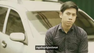 Young man with a serious expression sits beside a car, with text overlay in an Asian language at the bottom of the image.