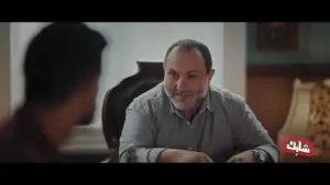 A smiling man seated at a table converses with another person in a cozy, well-lit room.