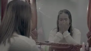 A distressed woman with long hair covers her face while looking into a mirror, capturing an emotional moment.