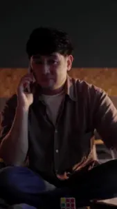 Man sitting cross-legged on the floor, talking on the phone in a dimly lit room, with a Rubik's Cube nearby.