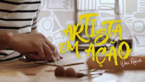 Creative artist crafting handmade items, with vibrant yellow text overlay reading "Artista em Ação" in a studio setting.