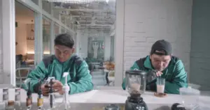 Two young men in green jackets sit at a cafe, one texting and the other sipping a drink, with a modern interior in the background.