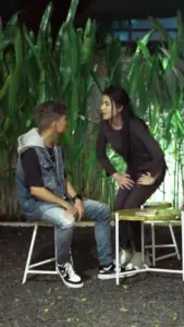 A young man and woman engage in a lively conversation, seated on stools surrounded by lush greenery.