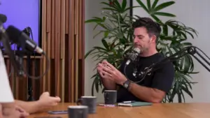 Podcast hosts discussing topics at a table surrounded by plants and mugs, with microphones set up for recording.