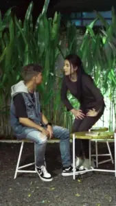 A young man and woman engage in a lively conversation amidst lush greenery at night, creating an intimate atmosphere.