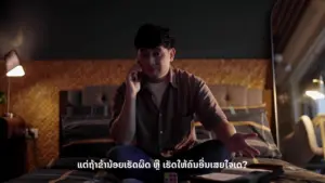 Young man sitting on a bed, talking on the phone, surrounded by a cozy bedroom atmosphere in soft lighting.