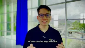 Man with glasses speaking enthusiastically in front of large windows, wearing a dark shirt with a logo.