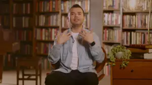 Man seated in a cozy library, gesturing expressively with books and plants in the background, conveying a thoughtful moment.