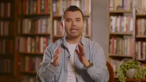 Man gesturing while seated in a cozy library, surrounded by bookshelves filled with colorful books.