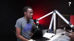 Young man with glasses speaking into a microphone in a modern radio studio with red and black backdrop and computer setup.