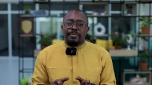 Man in a yellow shirt with glasses stands in front of a green plant backdrop, giving an engaging presentation.