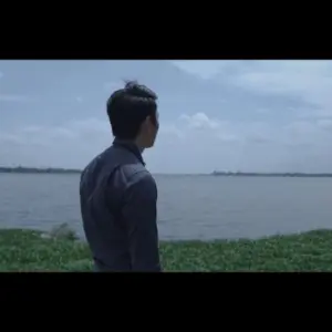 Man in a blue shirt gazing at the serene river under a cloudy sky, surrounded by lush greenery.