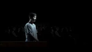 A young boy stands alone under dramatic lighting in a darkened room, surrounded by an audience in shadow.