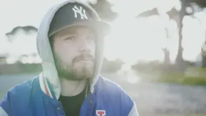 Young man in a hoodie and cap stands outdoors, with sunlight glowing in the background, creating a soft, dreamy atmosphere.