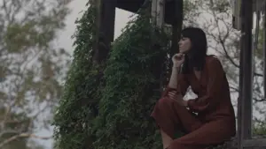 Woman in a rust-colored outfit sits thoughtfully among lush greenery, reflecting on nature and tranquility.