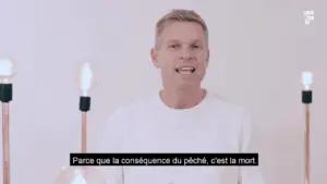 Man in a white sweater speaking in front of light bulbs, discussing a serious topic with subtitles in French.