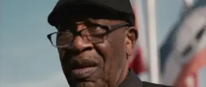 Elderly man wearing glasses and a black cap, expressing deep emotions against a backdrop of flags.