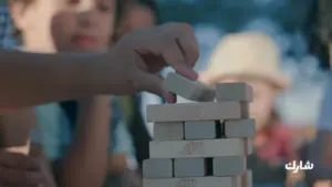 Child's hand stacking a wooden block in a game, with friends watching in the background on a sunny day outdoors.