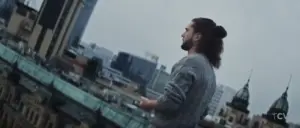 Man with a ponytail standing on a rooftop, overlooking a city skyline on a cloudy day.