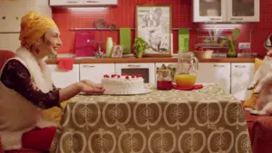 Woman in a kitchen smiles while holding a cake on a table set with drinks, showcasing a cheerful celebration scene.