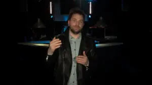 Man in a leather jacket gestures on stage with blue lights and musical equipment in the background, creating a dynamic atmosphere.