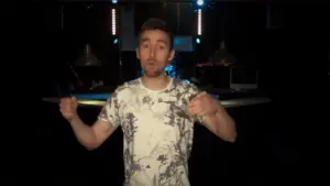 Young man in a floral shirt gestures confidently in a vibrant stage setting with lights and equipment in the background.