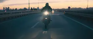 A motorcyclist riding on a highway at sunset, with city skyline in the background and headlights illuminating the road.