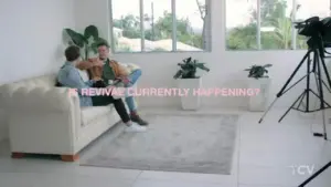 Two people sit on a white couch in a bright room discussing a topic, with plants and a camera visible in the background.