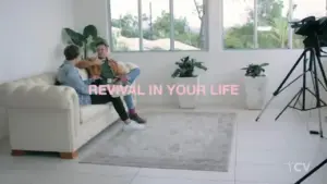 Two people seated on a couch in a bright room, discussing topics of personal growth with a camera nearby.