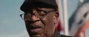 Elderly man wearing glasses and a black cap, with flags in the background, expressing a thoughtful demeanor.