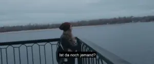 A person in a red beanie stands by a railing, gazing at a calm river under a cloudy sky.