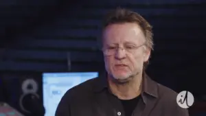 A middle-aged man with glasses discusses music production in a studio setting, with electronic equipment in the background.