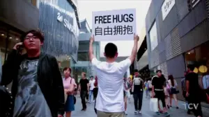 A person holding a "Free Hugs" sign in a busy urban area, surrounded by a diverse crowd of pedestrians.