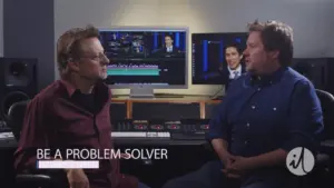 Two men discussing video editing in a modern studio, with editing software and a screen showing a presenter in the background.