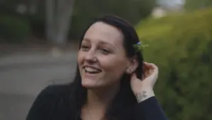 Woman smiling outdoors with a flower tucked behind her ear, surrounded by greenery and a blurred background.