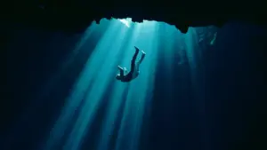 Person diving underwater with beams of light illuminating the scene from above in a serene blue environment.