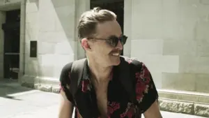 Man in a floral shirt and sunglasses smiles while walking on a sunlit street, exuding a relaxed, stylish vibe.