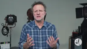 Man in plaid shirt gesturing expressively in a studio setting with film equipment in the background.