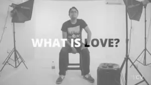 Man sitting on a chair in a studio setting, contemplating the question "What is love?" with dramatic lighting.