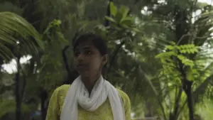 A young woman in a yellow outfit and white scarf stands against a lush green background of trees and foliage.