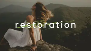 Woman in a white dress on a rock, enjoying nature with the word "restoration" overlayed against a mountainous backdrop.