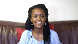 Smiling woman with braided hair sitting on a sofa, wearing a light blue top, against a neutral background.